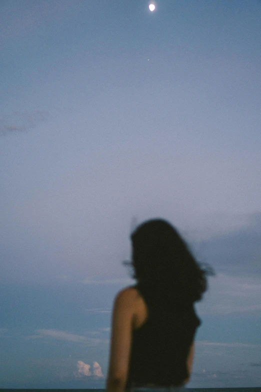 a woman standing on top of a beach next to the ocean, a picture, unsplash, conceptual art, windblown dark hair, evening sky, photo taken on fujifilm superia, blurred