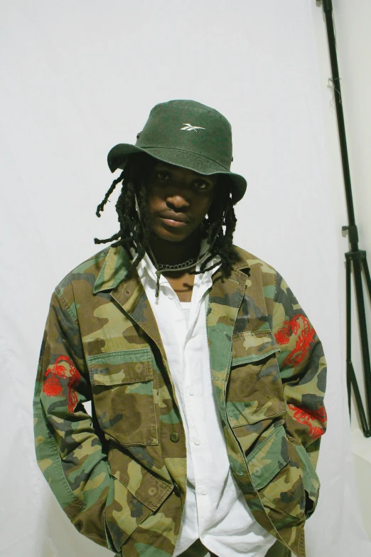 a man with dreadlocks standing in front of a white backdrop, bucket hat, ((zerator)), wearing military outfit, playboi carti