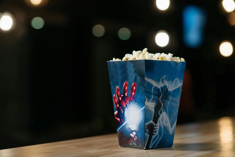 a popcorn box sitting on top of a wooden table, a hologram, avengers, back of hand on the table, at night, arthouse cinema