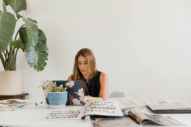 a woman sitting at a table working on a laptop, a picture, by Nicolette Macnamara, trending on unsplash, in a white room, magazine photo, 9 9 designs, flowers around