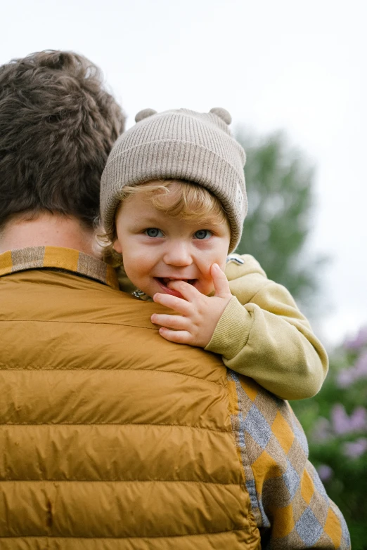 a man holding a small child in his arms, pexels, hat and hoodie, model wears a puffer jacket, brown, zoomed in