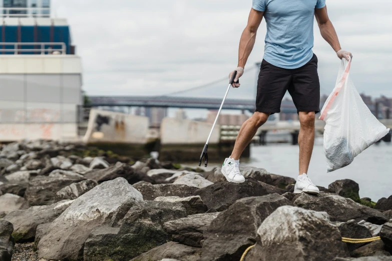 a man standing on top of a pile of rocks, wearing golf shorts, stepping on the city, holding a cane, standing next to water