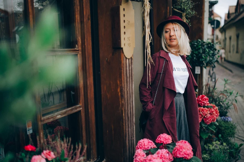 a woman standing in front of a building with flowers, by Julia Pishtar, pexels contest winner, maroon hat, wearing a trenchcoat, standing in a restaurant, gandalf as a woman