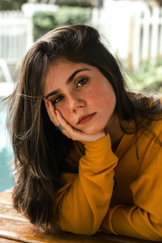 a woman sitting on top of a wooden table next to a pool, inspired by Elsa Bleda, trending on pexels, photorealism, wearing a yellow hoodie, portrait of depressed teen, brown hair and large eyes, maya ali