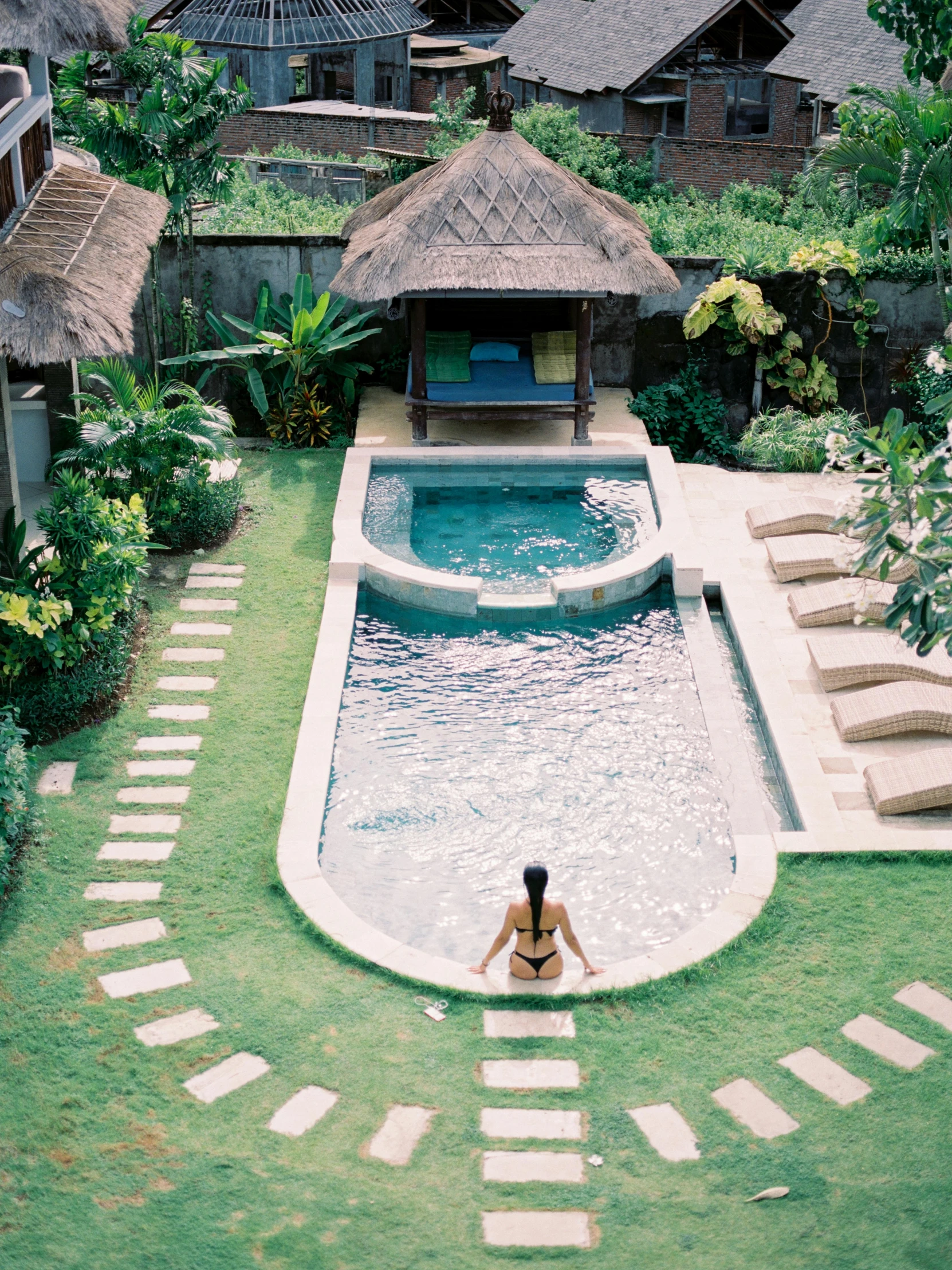 a woman sitting at the edge of a swimming pool, above lush garden and hot spring, bird view, nostalgic vibes, villa