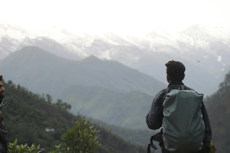 a couple of men standing on top of a lush green hillside, inspired by Sunil Das, pexels contest winner, a man wearing a backpack, man sitting facing away, snow capped mountains, looking across the shoulder