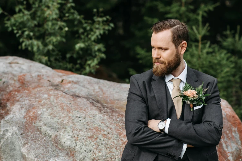 a man in a suit and tie standing in front of a rock, by Jaakko Mattila, unsplash, wedding, well trimmed beard, flowers around, “ iron bark