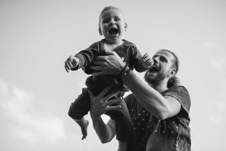 a man holding a baby up in the air, a black and white photo, by Matija Jama, pexels, figuration libre, laughing hysterically, avatar image, ryan church, enjoying the wind