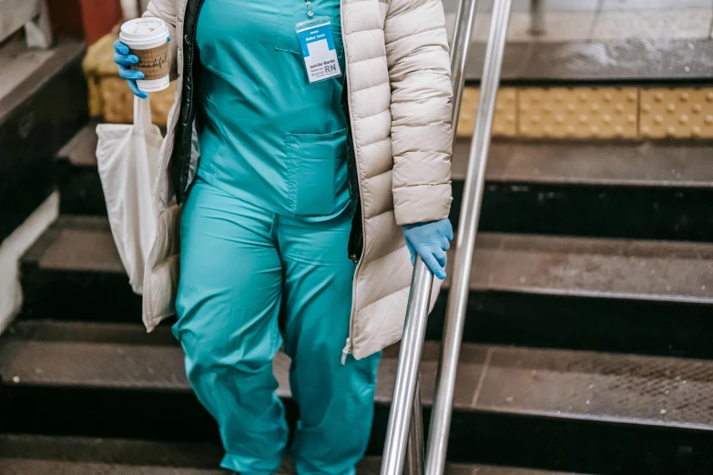 a woman walking down a flight of stairs holding a cup of coffee, surgical gown and scrubs on, wearing teal beanie, reflective suit, iv drip