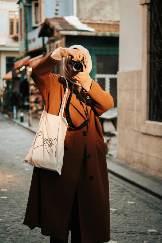 a woman taking a picture of a street with a camera, by irakli nadar, pexels contest winner, wearing a long beige trench coat, bags, hijab, cartoonist