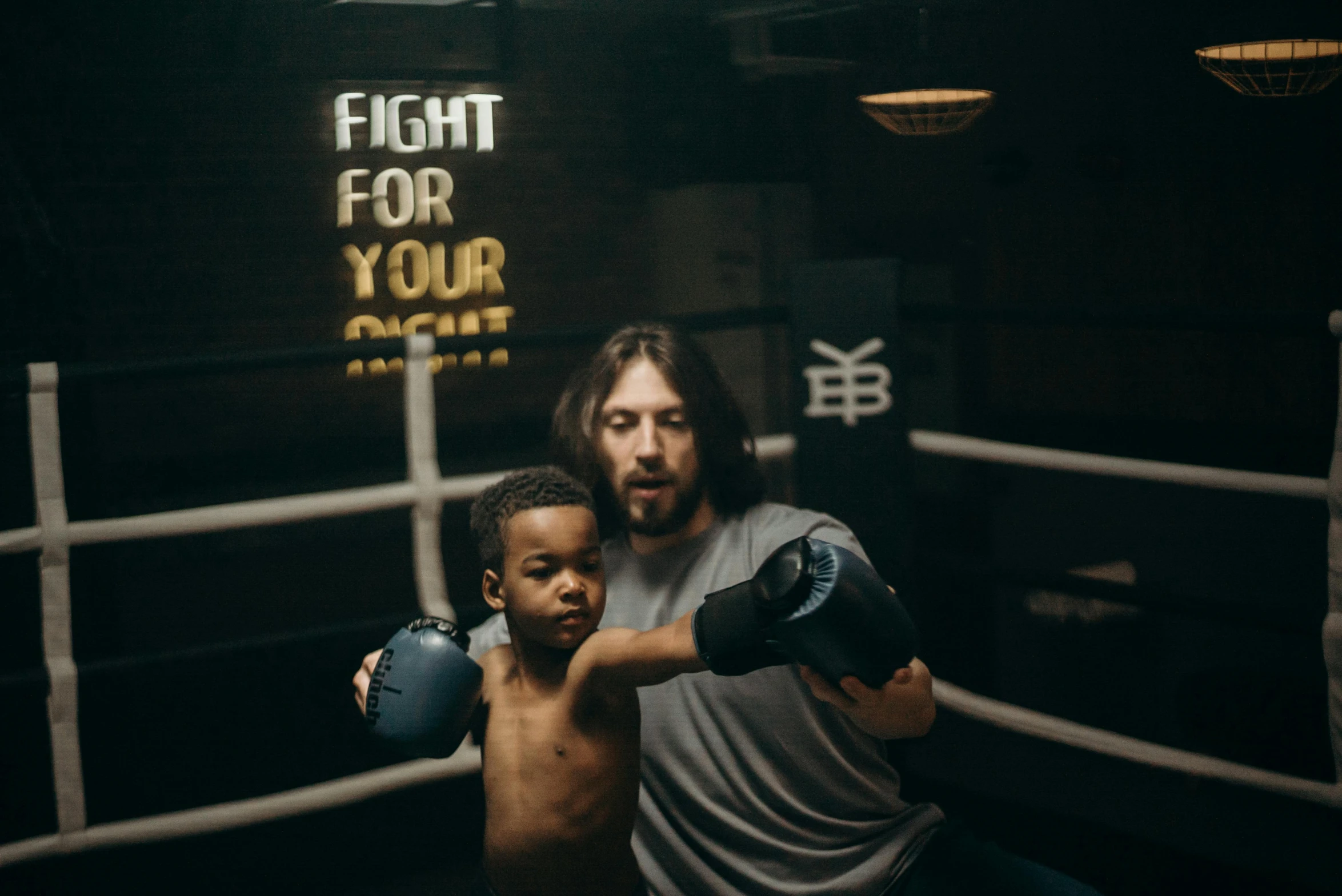 a man standing next to a young boy in a boxing ring, an album cover, inspired by Jeremy Henderson, pexels contest winner, dave grohl, lit up in a dark room, action with run and fight, bowater charlie and brom gerald