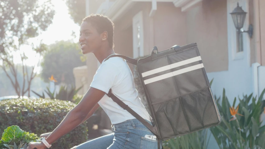 a woman riding a bike with a bag on the back, happening, carrying a tray, profile image, square backpack, dark-skinned