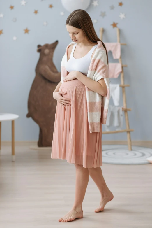 a pregnant woman standing in a room next to a teddy bear, shutterstock, renaissance, pleated skirt, in shades of peach, wearing skirt, warmer and softer colours