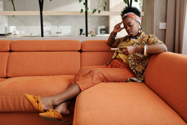 a woman sitting on a couch using a cell phone, by Lily Delissa Joseph, trending on pexels, afrofuturism, orange jumpsuit, topknot, old couch, mid-century modern furniture