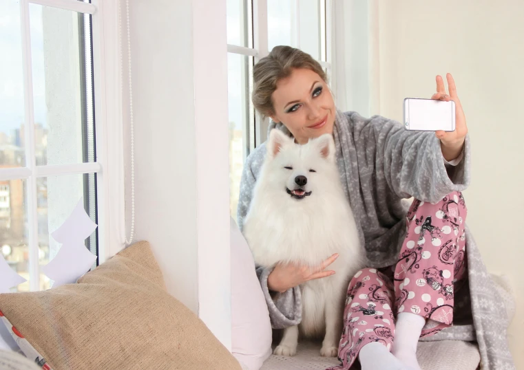 a woman taking a selfie with her dog, a picture, wearing pajamas, pomeranian mix, white, cozy calm! fabrics textiles