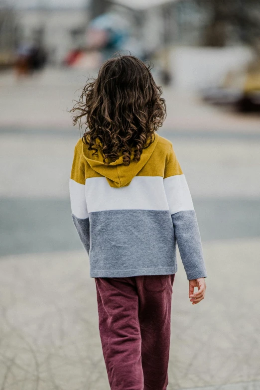 a little girl is walking down the street, inspired by Knox Martin, unsplash, solid colours material, with his back turned, wearing sweatshirt, warm tri - color