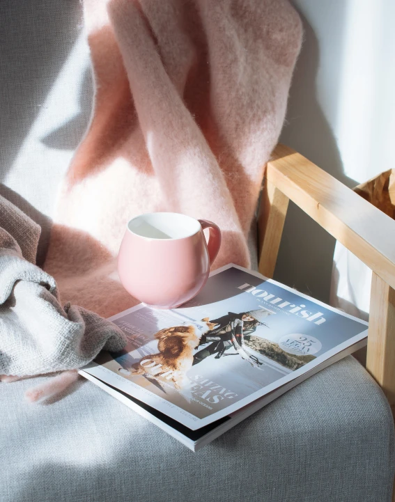 a book sitting on top of a chair next to a cup of coffee, a picture, white and pink cloth, magazine photograph, soft colours scheme, winter photograph