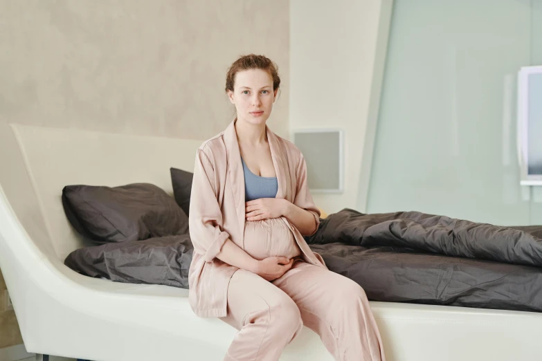 a pregnant woman sitting on a bed in a bedroom, by Nina Hamnett, pexels, happening, wearing a tracksuit, in a silk robe, ad image, nursing