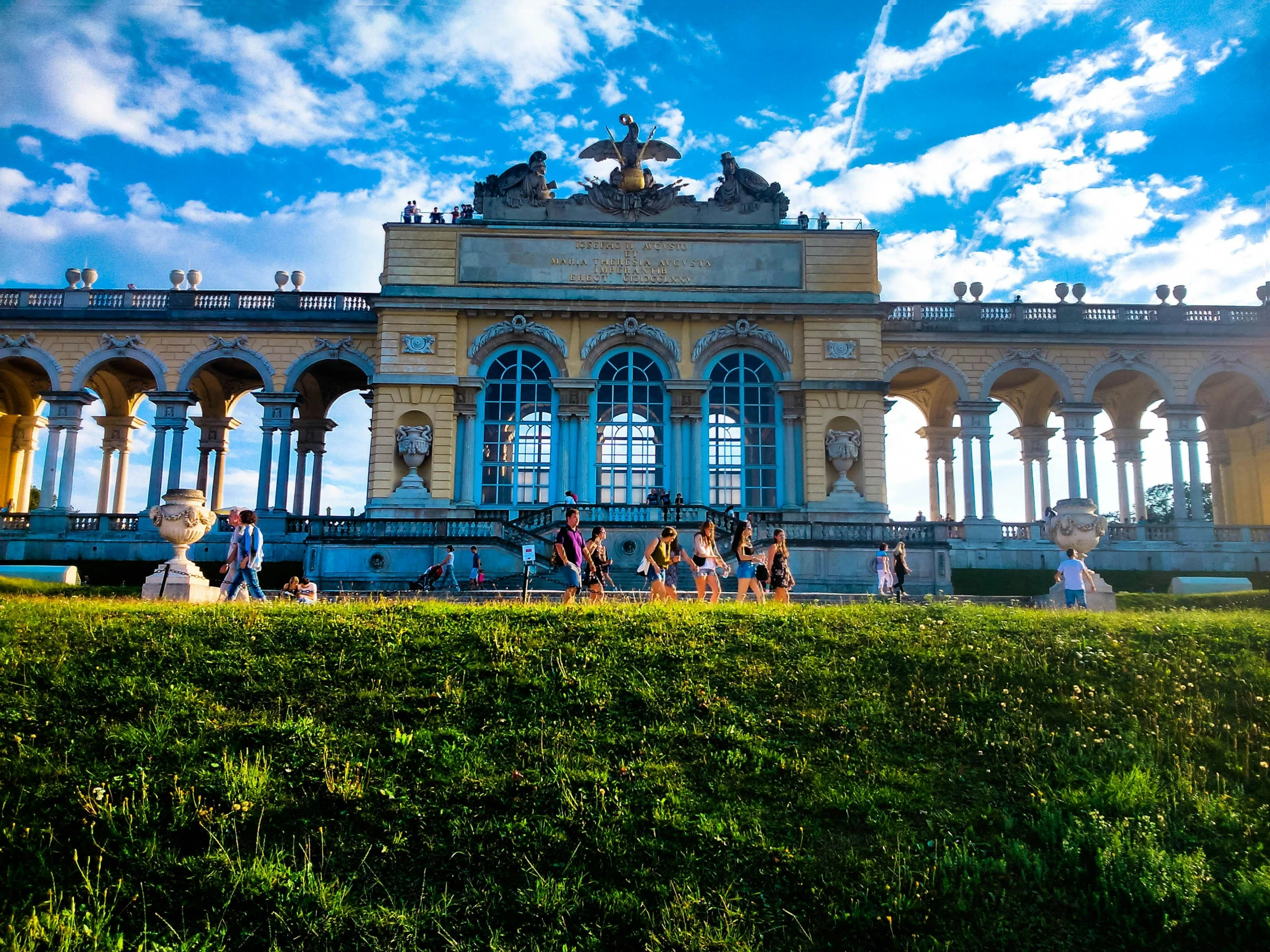 a large building sitting on top of a lush green field, an album cover, by Emanuel Büchel, pexels contest winner, neoclassicism, giant archways, people walking around, ornate with gold trimmings, fitness