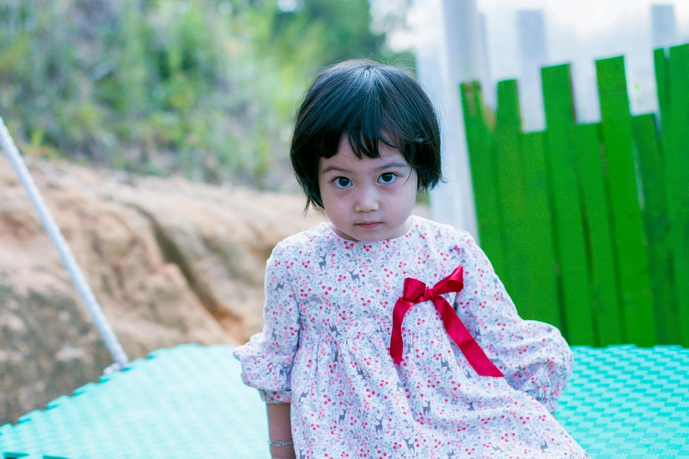 a little girl sitting on top of a green mat, a picture, inspired by Nazmi Ziya Güran, a red bow in her hair, taken with canon 8 0 d, but a stern look about her, in the garden