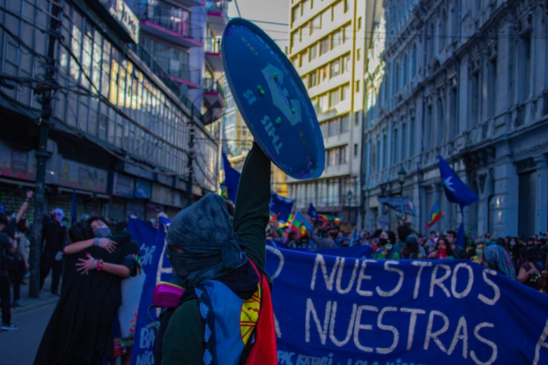 a large group of people walking down a street, by Luis Molinari, pexels contest winner, sots art, holding up a large shield, blue and violet, lady gaga president of argentina, subject action : holding sign