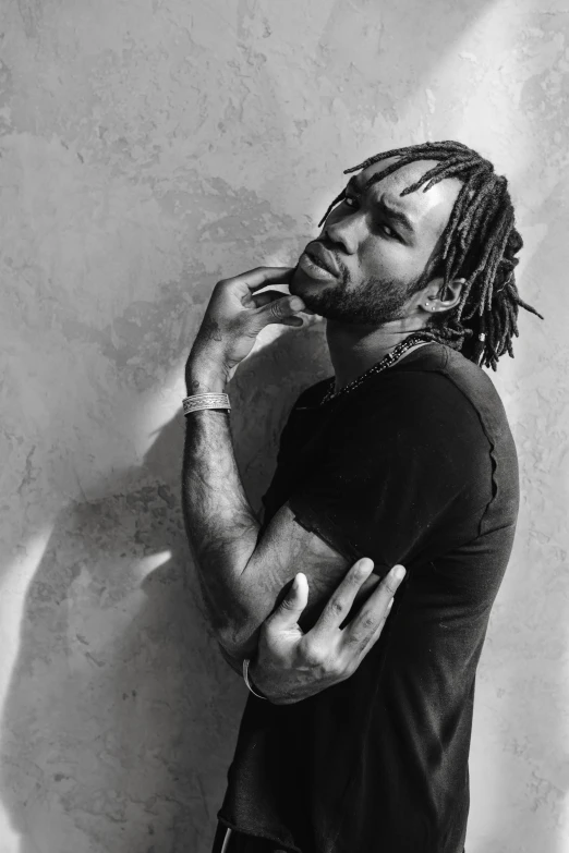 a man with dreadlocks leaning against a wall, a black and white photo, inspired by Richard Wright, playboi carti and lil uzi vert, he is wearing a black t-shirt, clasps his bangs in one hand, with textured hair and skin