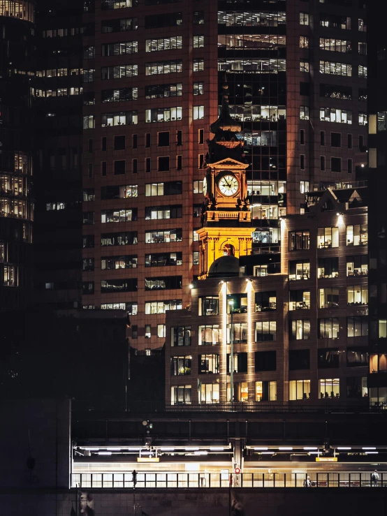 a clock tower in the middle of a city at night, a photo, inspired by George Pirie, australian tonalism, golden windows, full of high buildings, viewed from a distance, taken in the 2000s