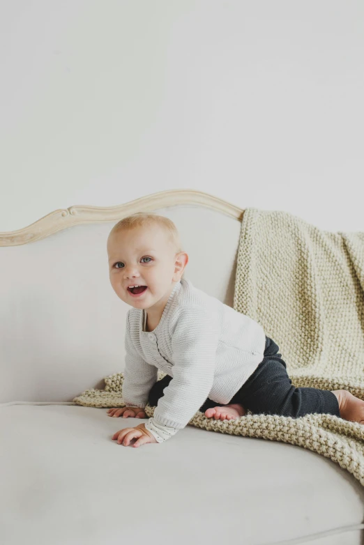 a baby sitting on top of a couch next to a blanket, inspired by Myles Birket Foster, unsplash, happening, neutral background, wearing a cardigan, smiling playfully, elegantly dressed
