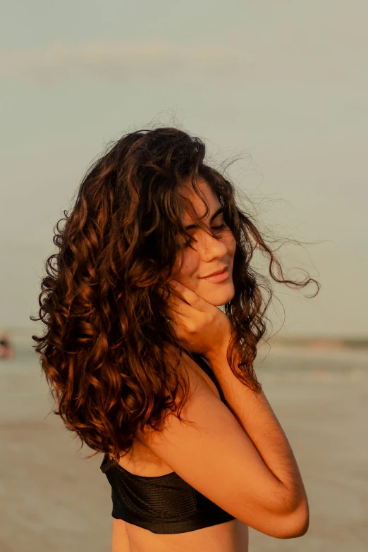 a woman in a black bikini standing on a beach, an album cover, pexels contest winner, curls hair, hands in her hair. side-view, at dusk at golden hour, long wavy brunette hair