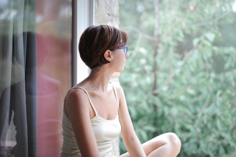 a woman sitting on a window sill looking out a window, natural short hair, anorexic figure, profile image, with glasses