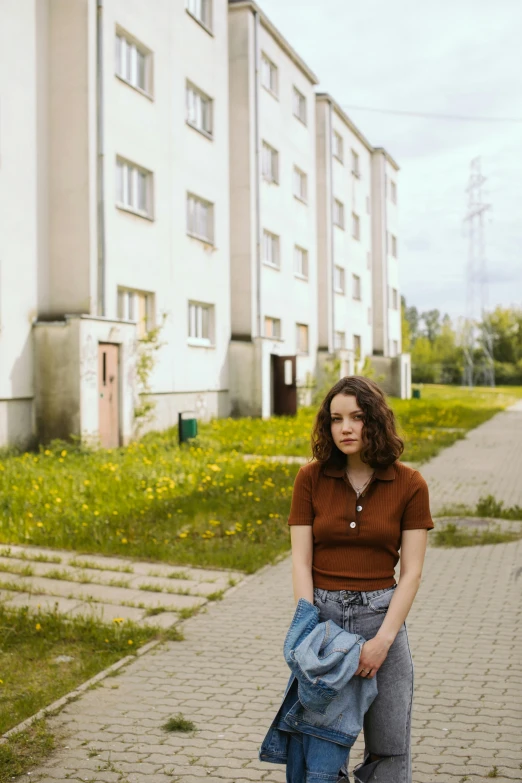 a woman standing on a sidewalk in front of a building, an album cover, inspired by Elsa Bleda, pexels contest winner, realism, soviet suburbs, standing in a grassy field, handsome girl, cynthwave