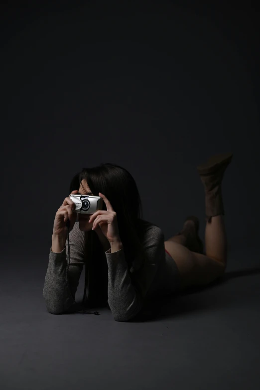 a woman laying on the ground holding a camera, art photography, dark backdrop, on grey background, sleek, joy ang