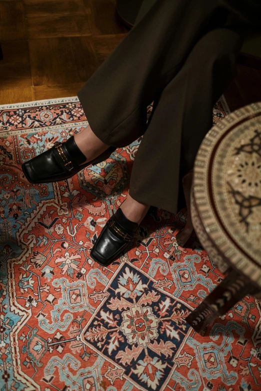 a woman sitting on top of a rug on top of a wooden floor, by Nina Hamnett, instagram, hurufiyya, black loafers, wes anderson and gucci, in a dark dusty parlor, ( ( theatrical ) )