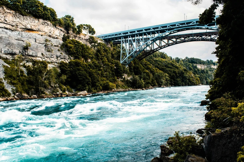 a bridge over a river next to a lush green forest, by Julia Pishtar, pexels contest winner, hurufiyya, niagara falls, clear blue water, vallejo, turquoise water