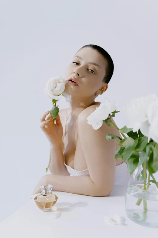 a woman sitting at a table with flowers in front of her, an album cover, inspired by Li Di, skincare, shaved hair, portrait sophie mudd, in white room