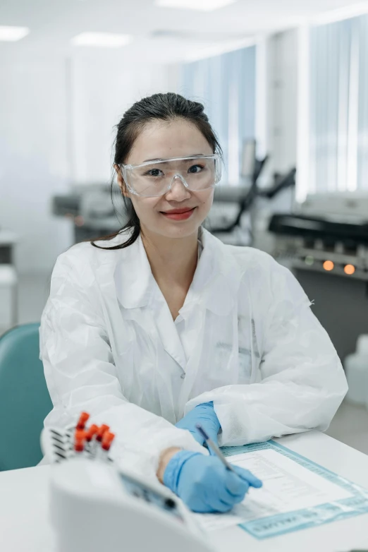 a woman in a lab coat and goggles sitting at a table, malaysian, confident looking, bio - chemical, profile image