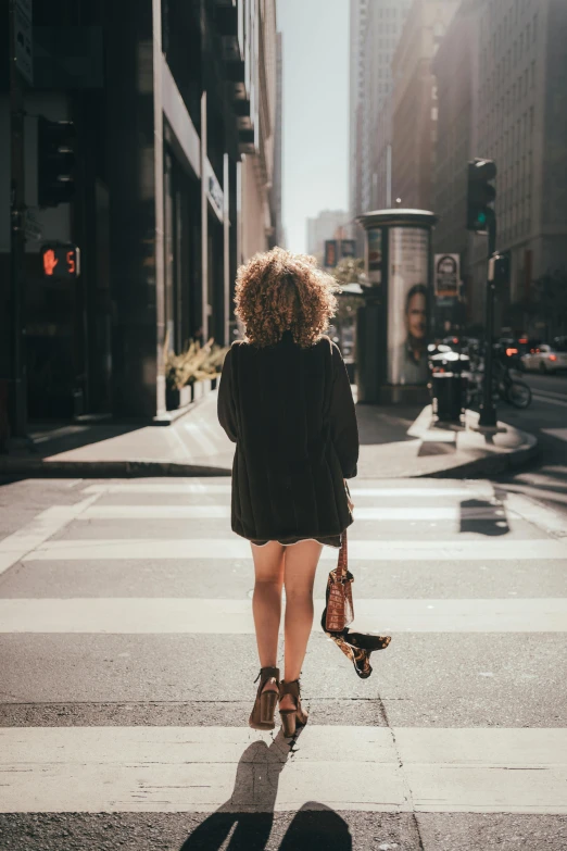 a woman walking across a street holding a skateboard, pexels contest winner, renaissance, curly haired, short skirt and a long jacket, facing away, african american young woman