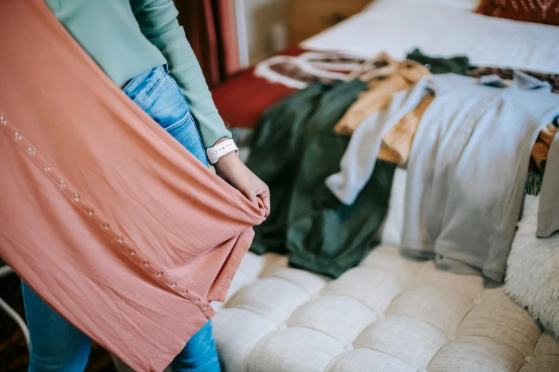 a woman standing in front of a bed holding a blanket, by Alice Mason, trending on unsplash, happening, green and brown clothes, pink clothes, exiting from a wardrobe, wide skirts