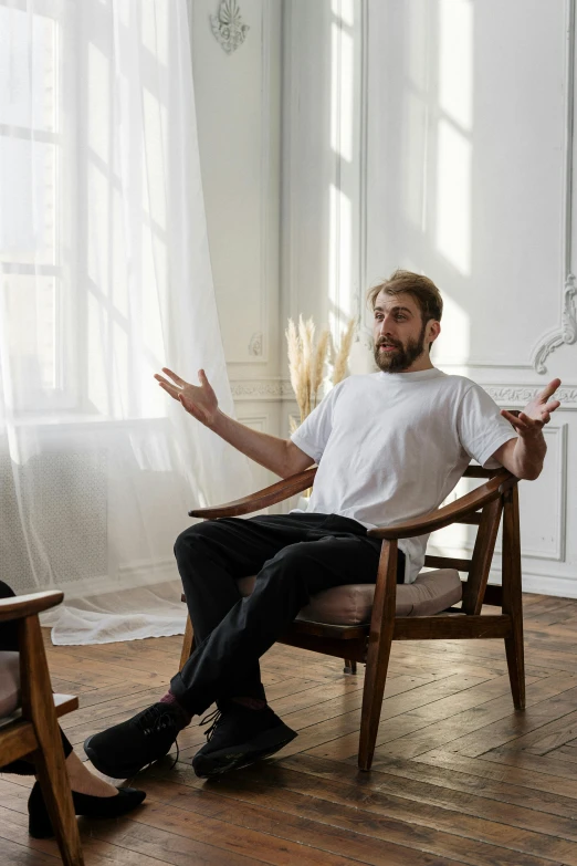 a man sitting in a chair in a room, inspired by Constantin Hansen, pexels contest winner, shrugging arms, bearded man, man in white t - shirt, ignant