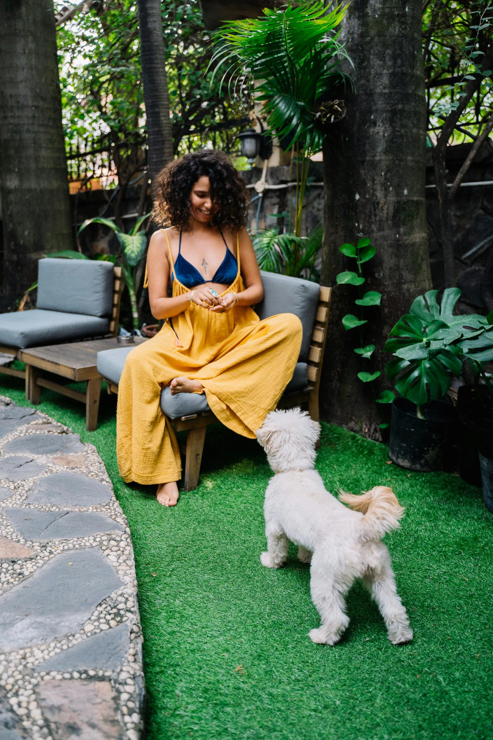 a woman sitting on a bench next to a dog, bali, wavy hair yellow theme, plants and patio, ultrawide image