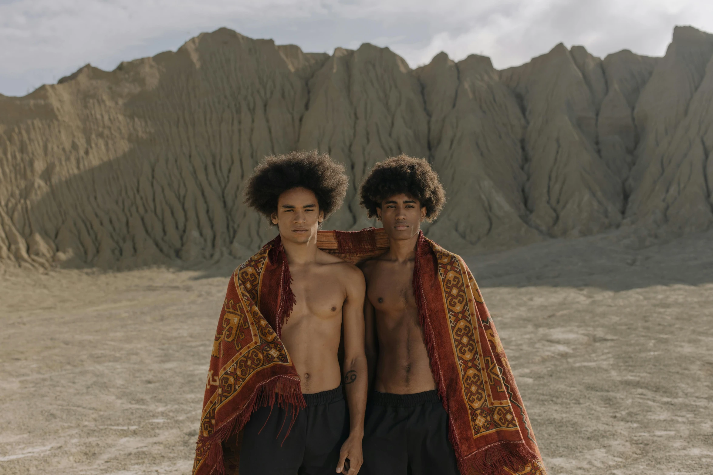 a couple of young men standing next to each other, by Alison Geissler, pexels contest winner, afrofuturism, desert robe, slightly tanned, with textured hair and skin, sarong