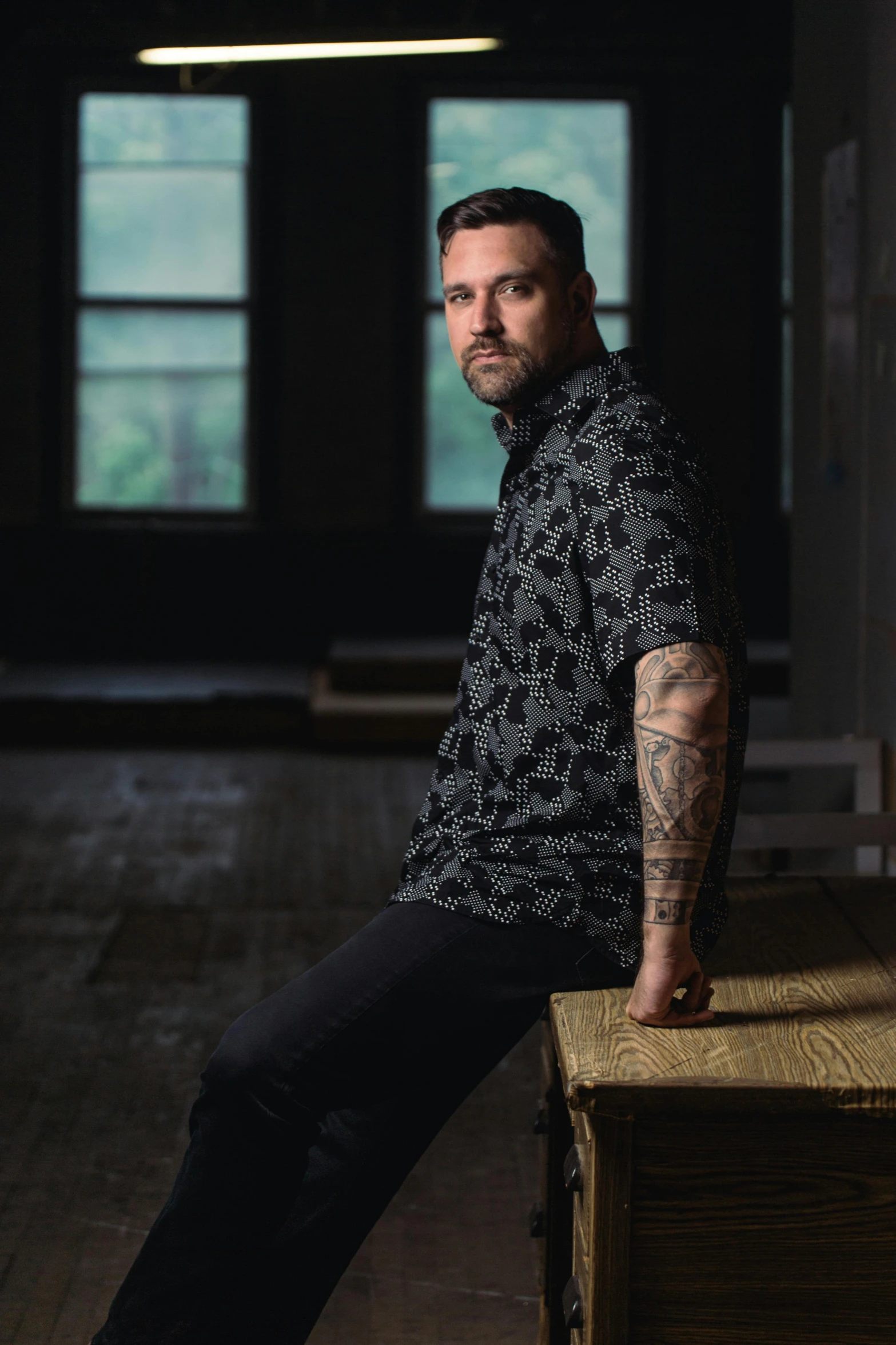 a man sitting on top of a wooden table, a portrait, by Jeremy Henderson, pexels contest winner, photorealism, tattooed, standing in a dimly lit room, paul thomas anderson, photograph of a sleeve tattoo