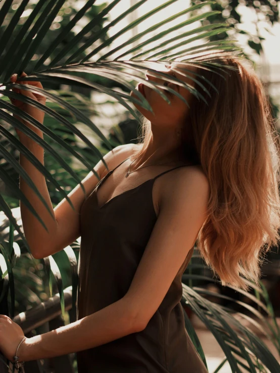 a woman standing next to a palm tree, inspired by Elsa Bleda, trending on unsplash, renaissance, light brown long wavy hair, on a planet of lush foliage, she is wearing a black tank top, hair : long brown