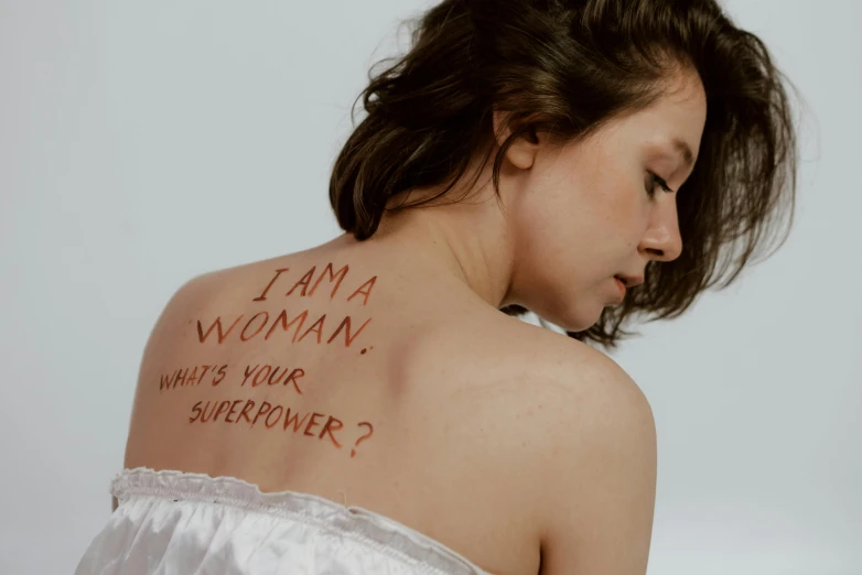 a woman with the words i am a woman written on her back, a tattoo, inspired by Tracey Emin, trending on pexels, feminist art, film still of gal gadot, superpowers, bodypainting, girl with brown hair