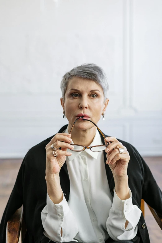 a woman sitting in a chair with a pen in her mouth, inspired by Ruth Deckard, trending on pexels, short grey hair, small square glasses, maurizio cattelan, mature