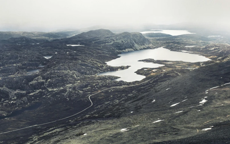 a man standing on top of a mountain next to a lake, by Jesper Knudsen, hurufiyya, aerial view of an ancient land, grey, dezeen, alpine tundra wildfire