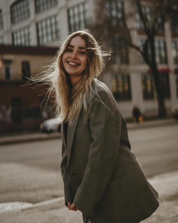 a woman standing in the middle of a street, trending on pexels, happening, grinning, grey jacket, non binary model, wind in hair