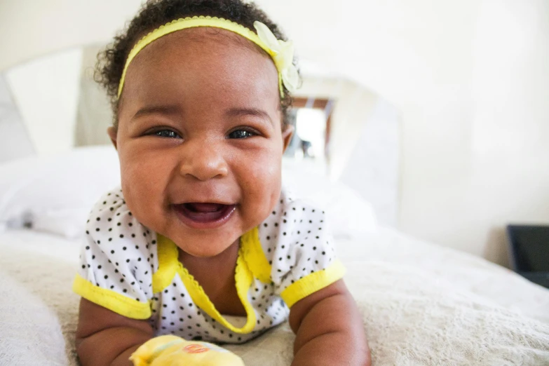 a close up of a baby laying on a bed, by Ella Guru, shutterstock contest winner, large black smile, lady using yellow dress, olive skinned, she has perfect white teeths