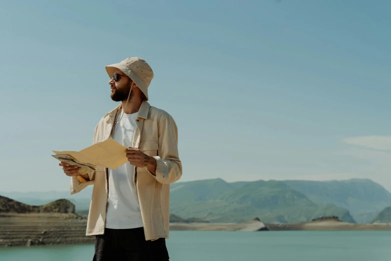 a man standing in front of a lake holding a piece of paper, an album cover, pexels contest winner, arab man light beard, straw hat and overcoat, standing in front of a mountain, streetwear