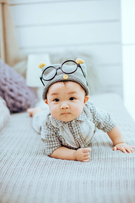 a baby laying on a bed wearing a hat and glasses, by Basuki Abdullah, happening, looking confident, square rimmed glasses, asian male, small